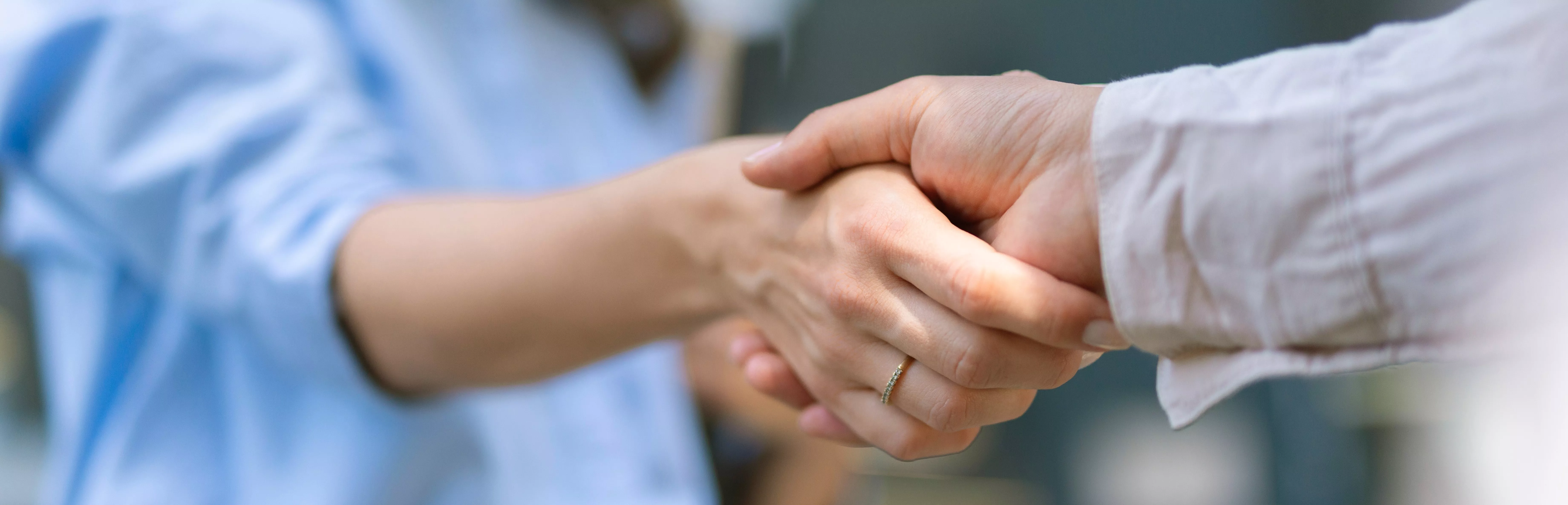Zwei Personen geben sich die Hand in einer Geste des Händeschüttelns, als Symbol für Zusammenarbeit. Eine Person trägt ein helles Hemd, die andere ein blaues Oberteil.